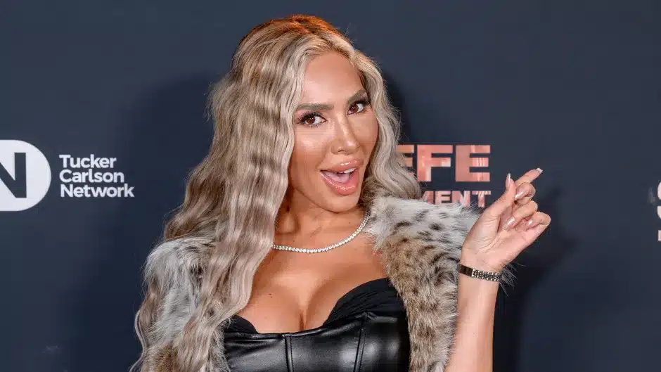 Person with long wavy hair poses in a fur-trimmed jacket and black top against a backdrop with logos, reminiscent of Farrah Abraham’s iconic style.
